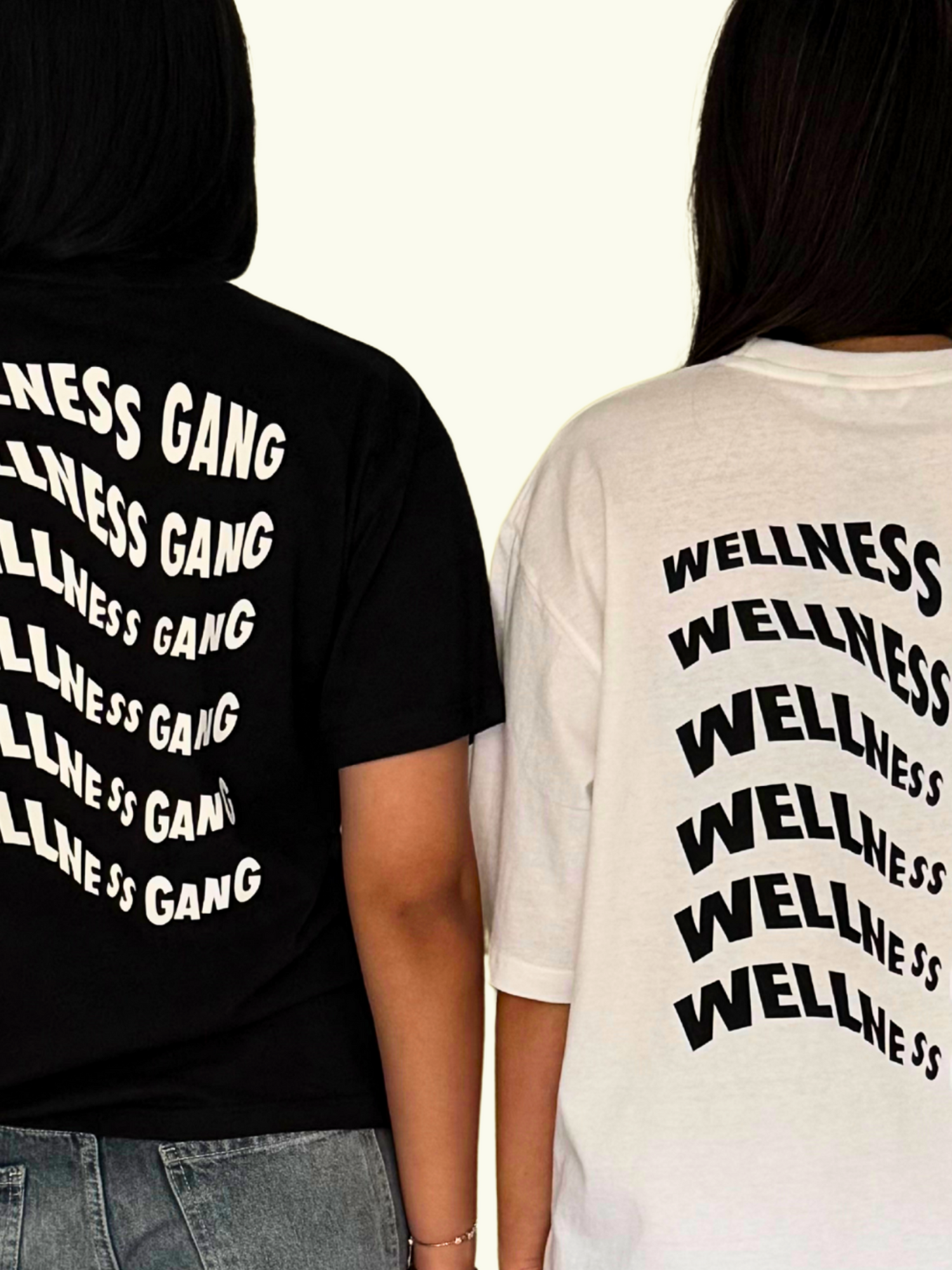 Two models wearing oversized t-shirts featuring 'Wellness Gang' graphics in black and white, showcasing organic cotton comfort.