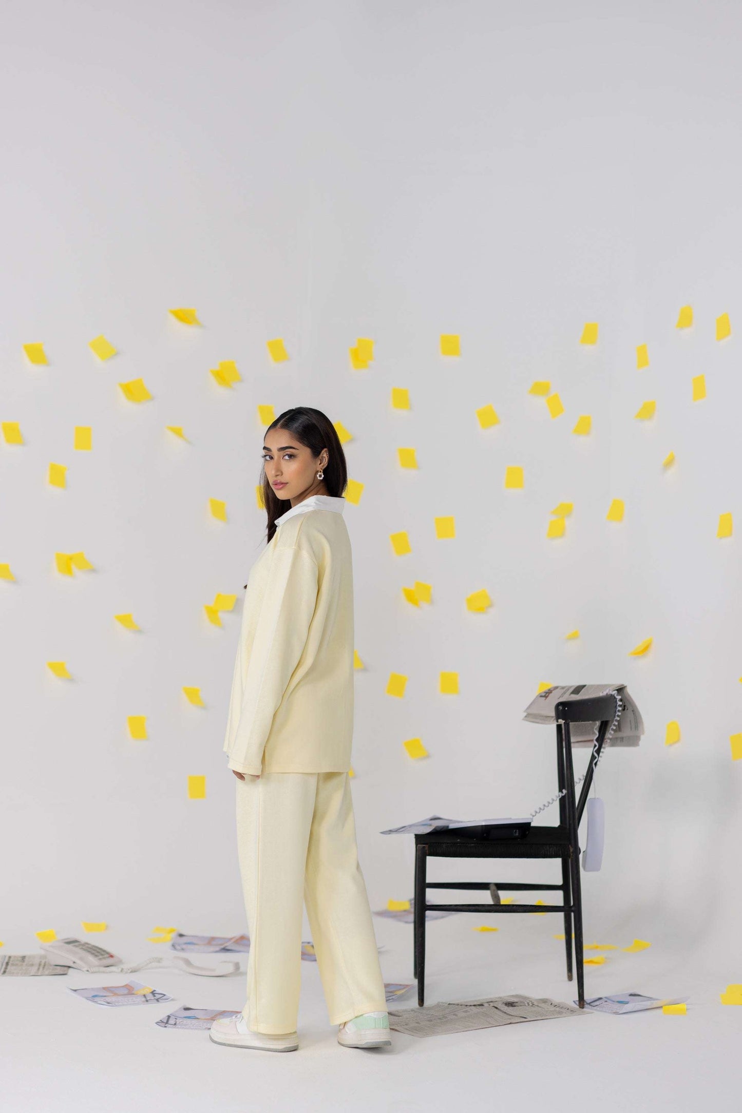 A model wearing cream oversized attire stands beside a black chair with yellow sticky notes floating around, showcasing a relaxed fit.