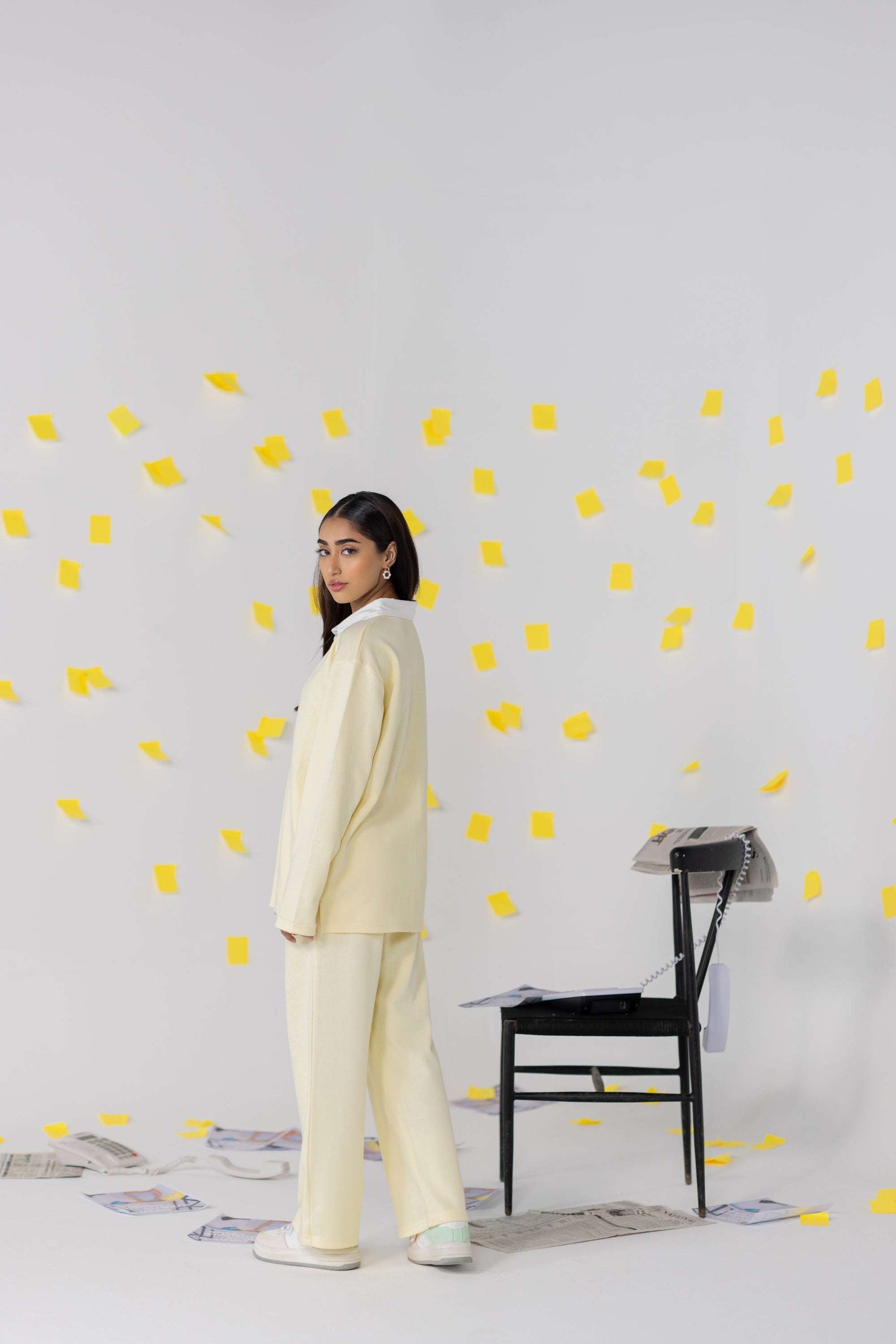 A model wearing cream oversized attire stands beside a black chair with yellow sticky notes floating around, showcasing a relaxed fit.
