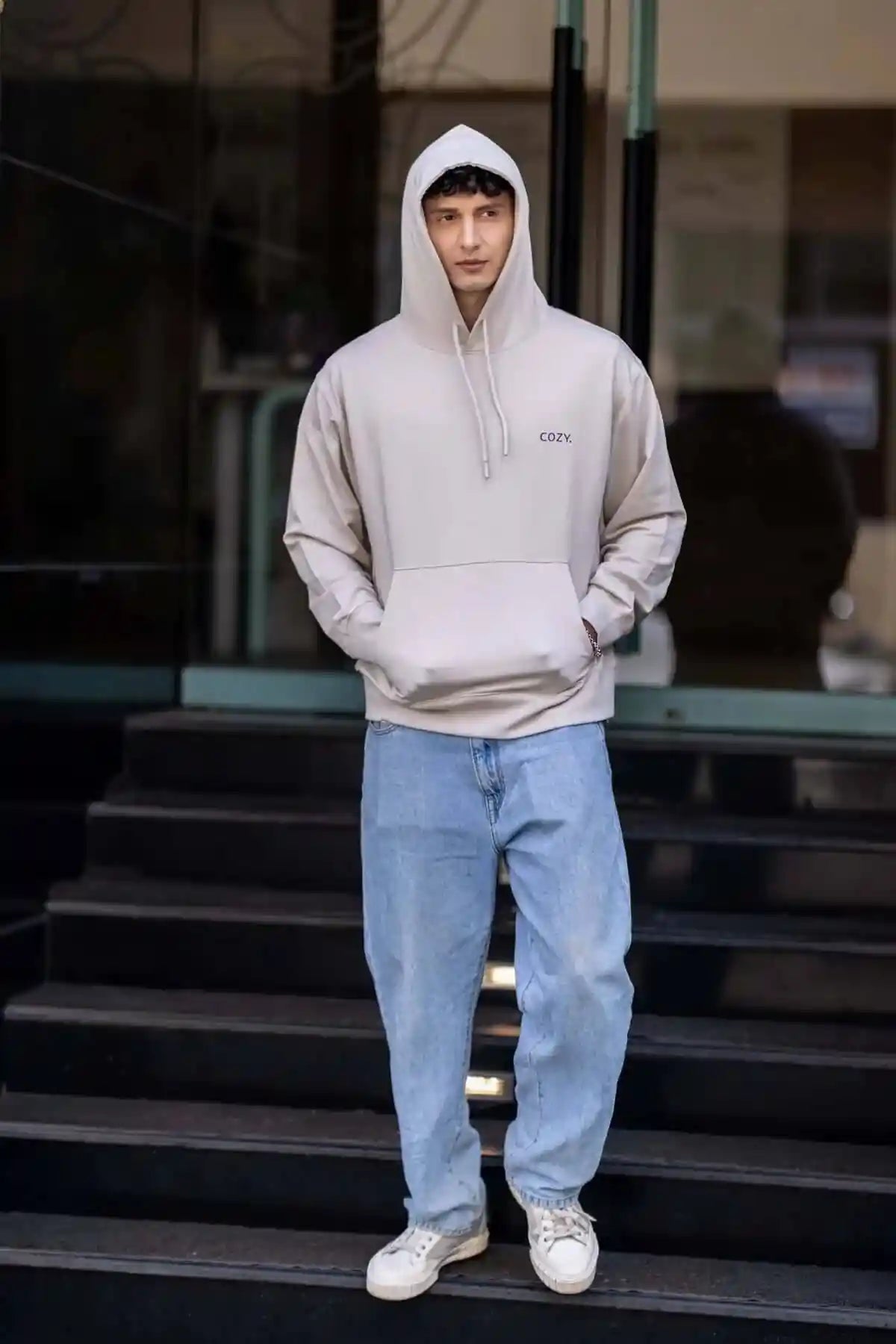 COZY Men's Hoodie in Cappuccino Beige worn hood up, hands in the kangaroo pocket, full length on dark stone steps
