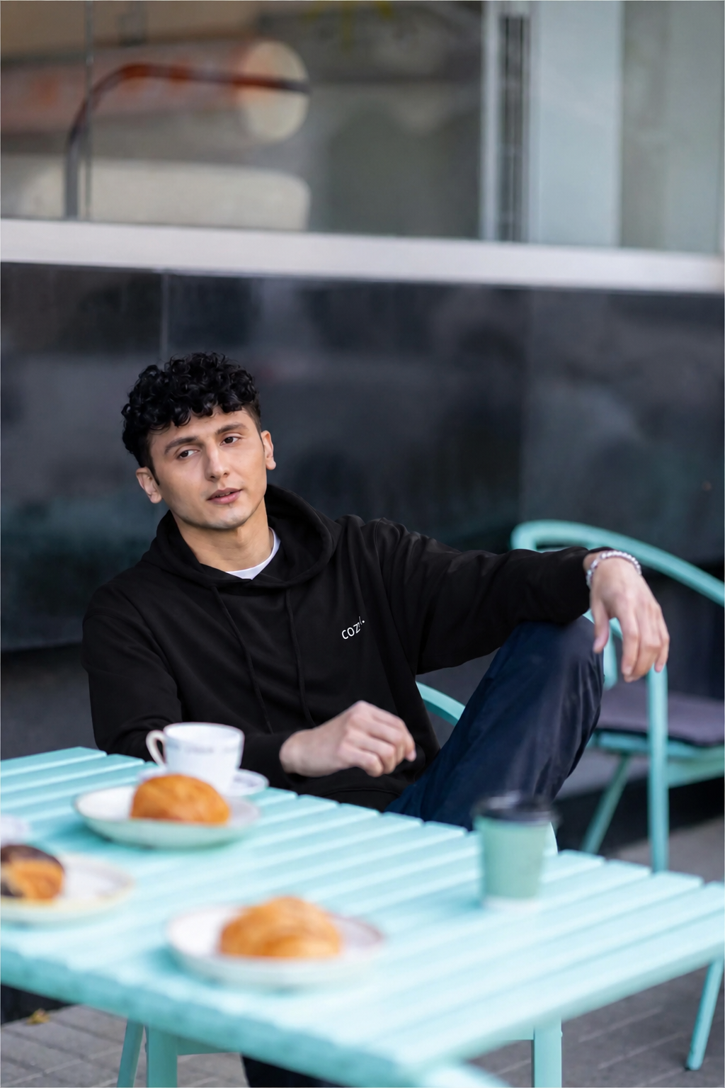 Man seated at a mint cafe table with coffee and croissants, wearing the COZY Men's Hoodie in Dark Roast Black