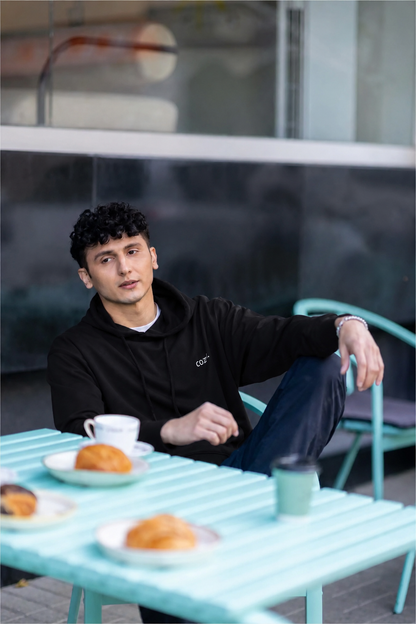 Man seated at a mint cafe table with coffee and croissants, wearing the COZY Men's Hoodie in Dark Roast Black