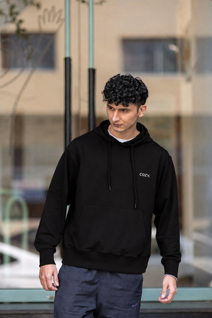 Front view of the COZY Men's Hoodie in Dark Roast Black, worn with dark trousers outside a glass storefront