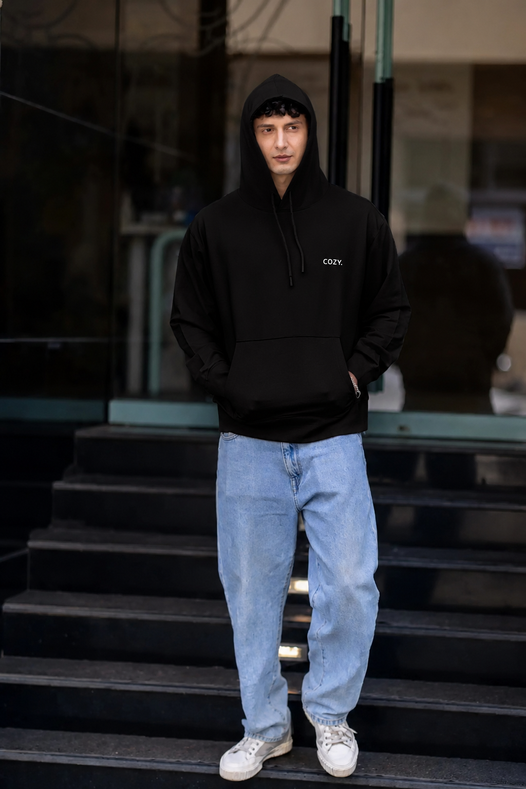 COZY Men's Hoodie in Dark Roast Black worn hood up with hands in the pocket, light wash jeans, full length on steps