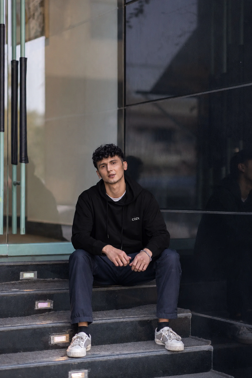 Man seated on stone steps wearing the COZY Men's Hoodie in Dark Roast Black with dark trousers and white sneakers