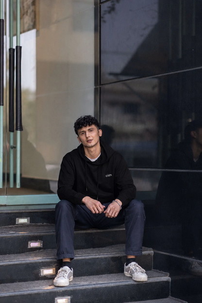 Man seated on stone steps wearing the COZY Men's Hoodie in Dark Roast Black with dark trousers and white sneakers