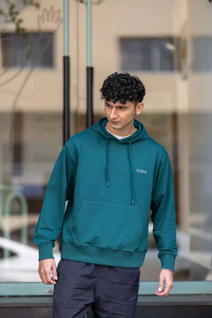 Front view of the COZY Men's Hoodie in Jade Brew, worn with dark trousers outside a glass storefront