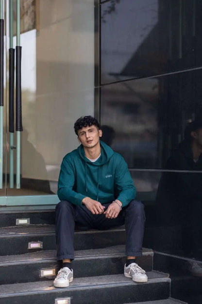 Man seated on stone steps wearing the COZY Men's Hoodie in Jade Brew with dark trousers and white sneakers
