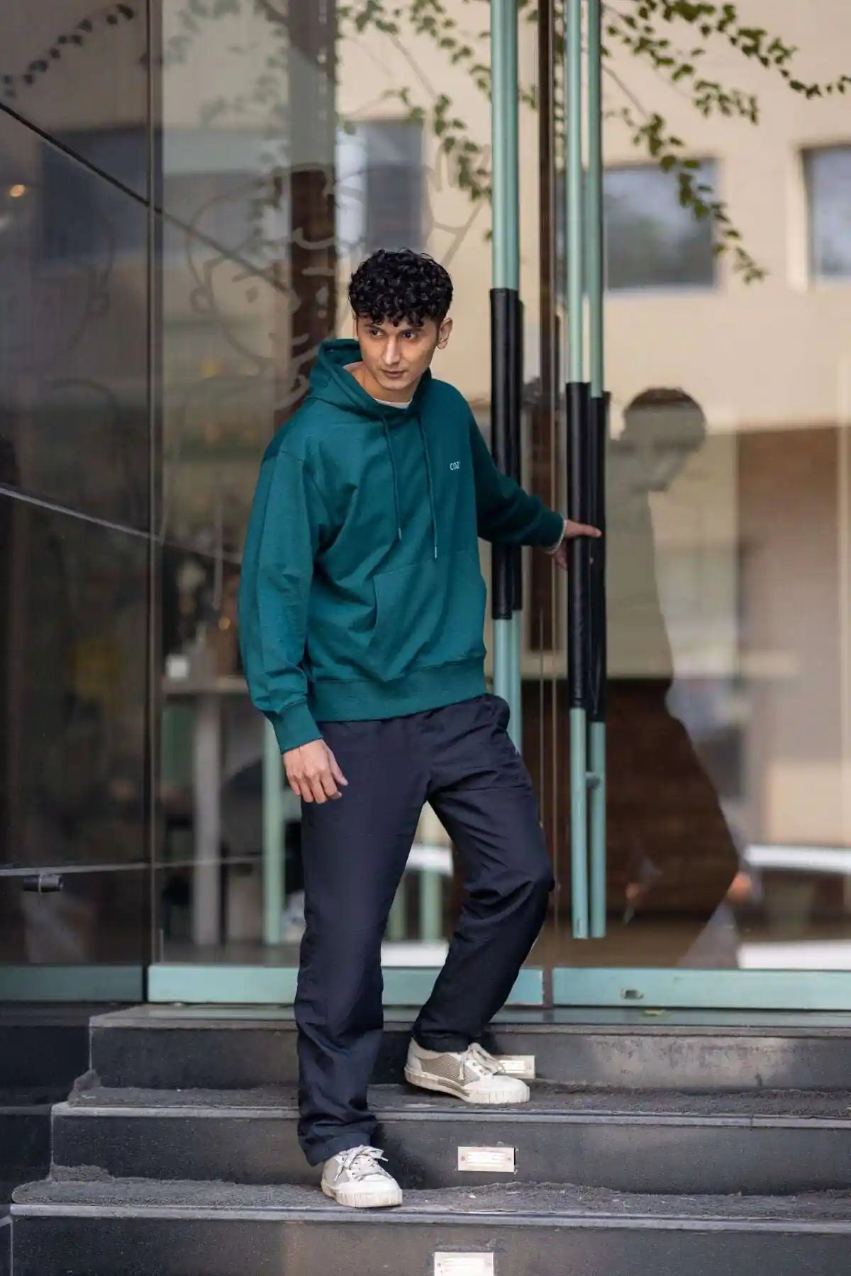 Side view of the COZY Men's Hoodie in Jade Brew, worn with dark trousers while stepping out of a glass door