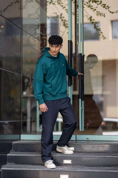 Side view of the COZY Men's Hoodie in Jade Brew, worn with dark trousers while stepping out of a glass door