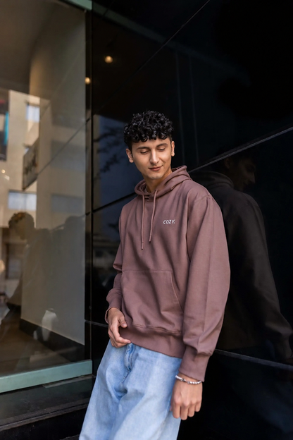 Man leaning against a dark glass wall in the COZY Men's Hoodie in Mocha Mousse, styled with light wash jeans