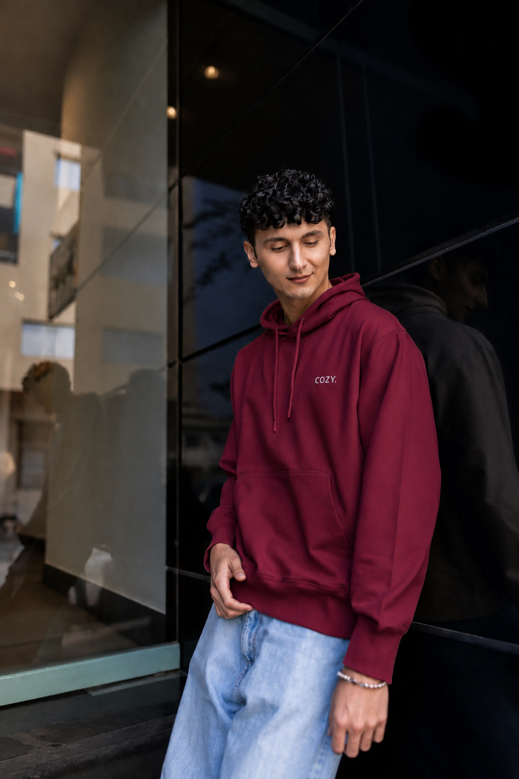 Man leaning against a dark glass wall in the COZY Men's Hoodie in Mulberry Mocha, styled with light wash jeans