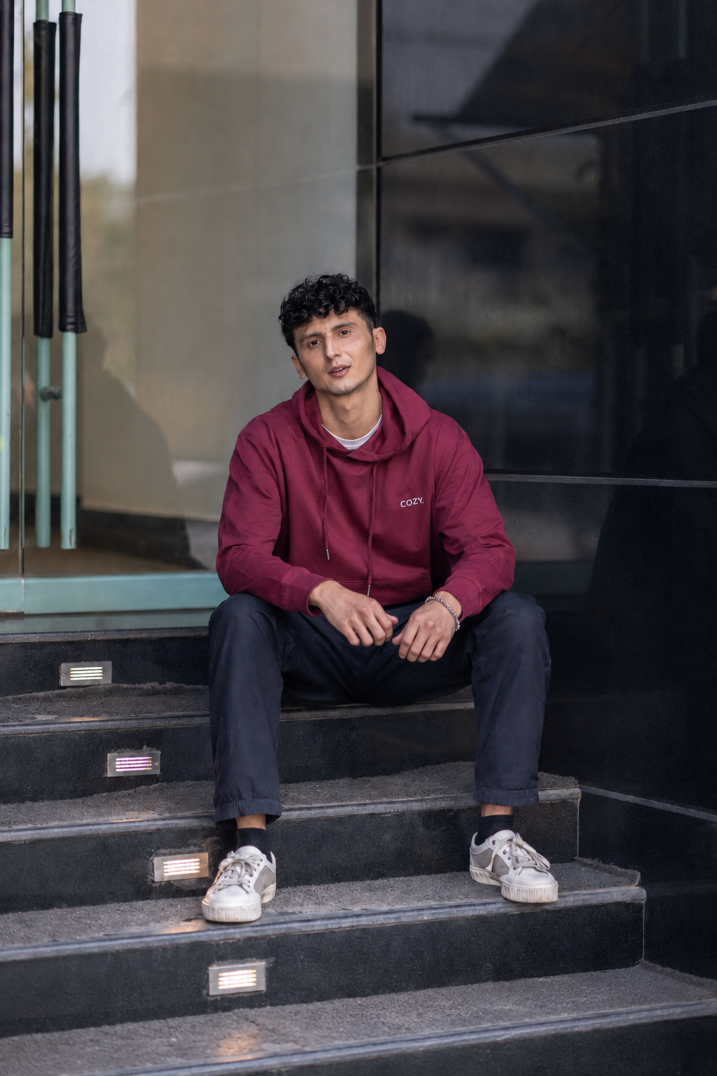 Man seated on stone steps wearing the COZY Men's Hoodie in Mulberry Mocha with dark trousers and white sneakers